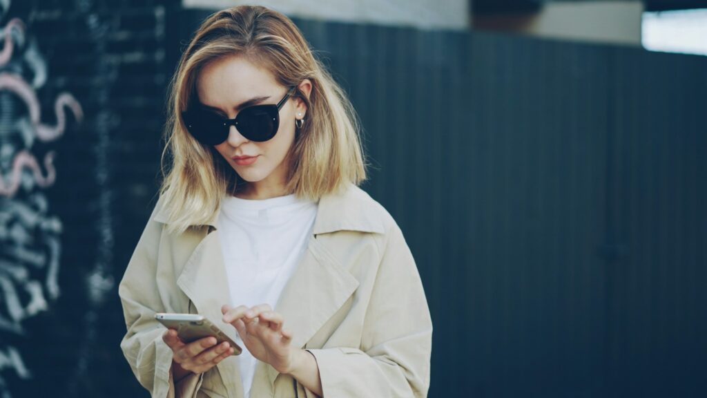 Vrouw met zonnebril gebruikt haar smartphone in een openbare ruimte, passend bij het thema mobiele etiquette.
